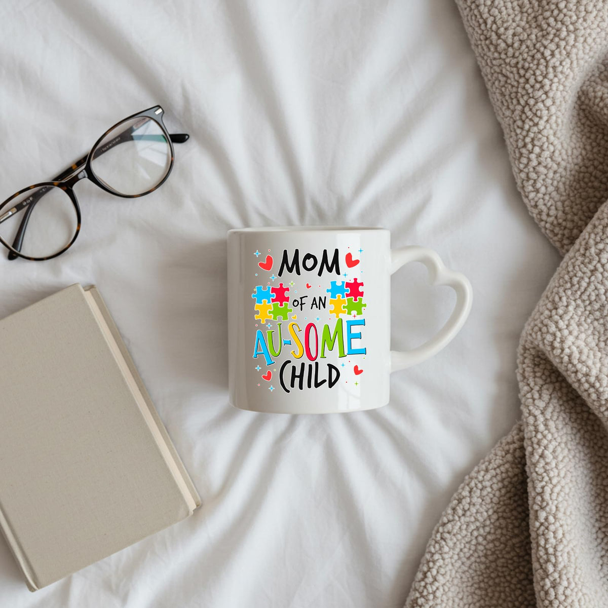 Mom of an Au-Some Child heart-handle mug on a bed beside glasses and a book