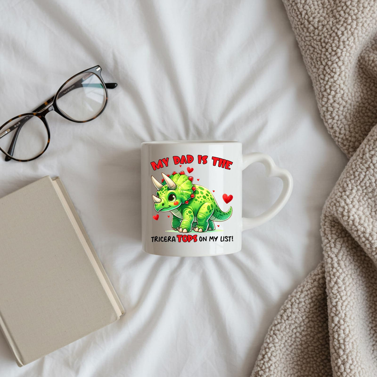 Triceratops dad heart-handle mug on a bed beside glasses and a book