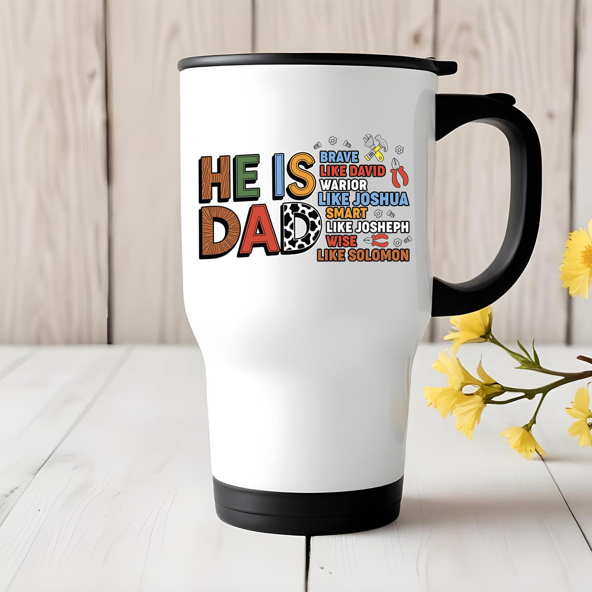 Travel mug with the He Is Dad Bible verse design on a white table with yellow flowers