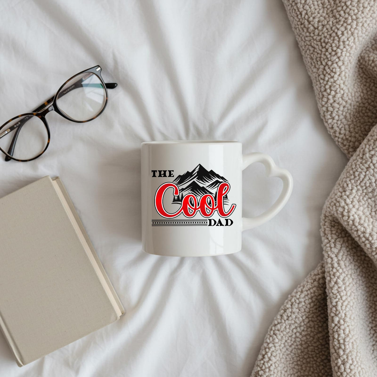 The Cool Dad mountains heart-handle mug on a bed beside glasses and a book