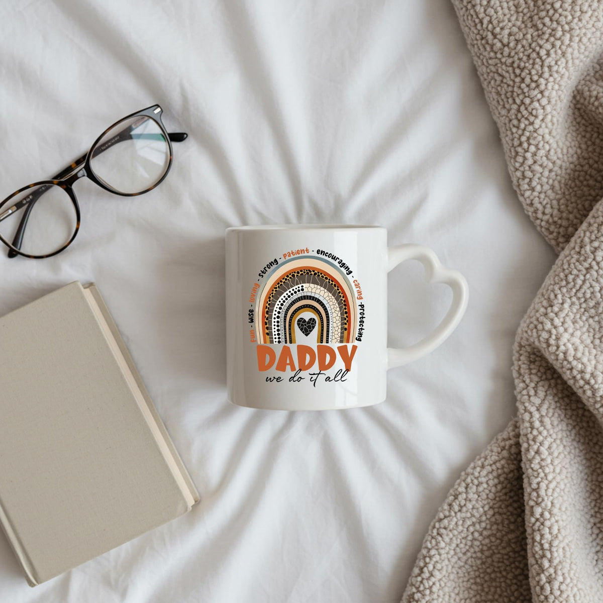 Daddy boho rainbow heart-handle mug on a bed beside glasses and a book