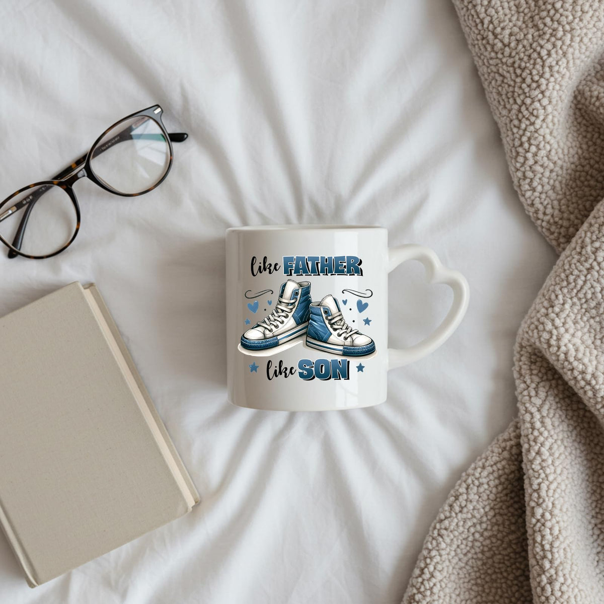 Like Father Like Son heart-handle mug on a bed beside glasses and a book