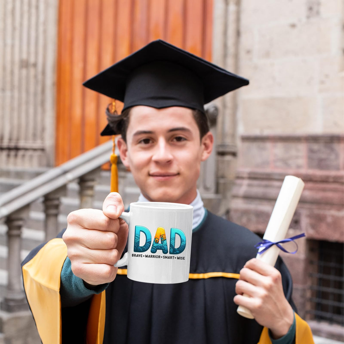 Graduate in cap and gown holding the DAD Brave Warrior mug