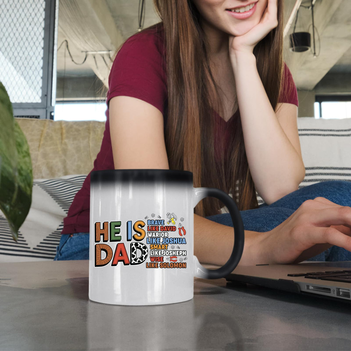 Woman at a laptop with a colour changing mug showing the He Is Dad Bible verse design