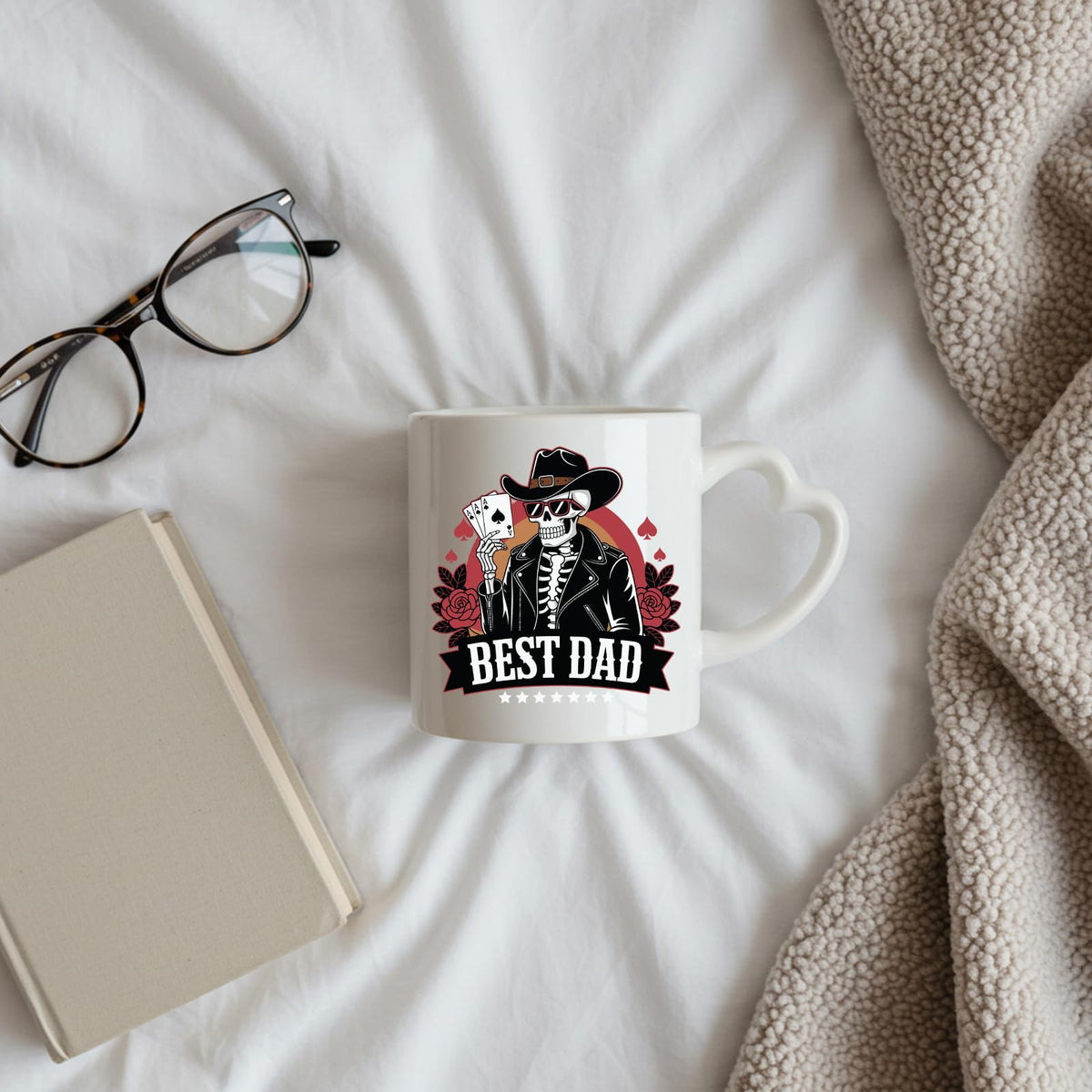 Best Dad skeleton cowboy heart-handle mug on a bed beside glasses and a book