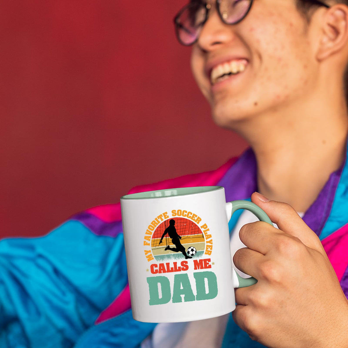 Smiling person in a colourful jacket holding the Soccer Dad mug with a light green handle