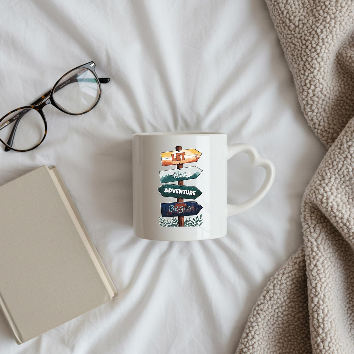 Let the Adventure Begin heart-handle mug on a bed beside glasses and a book
