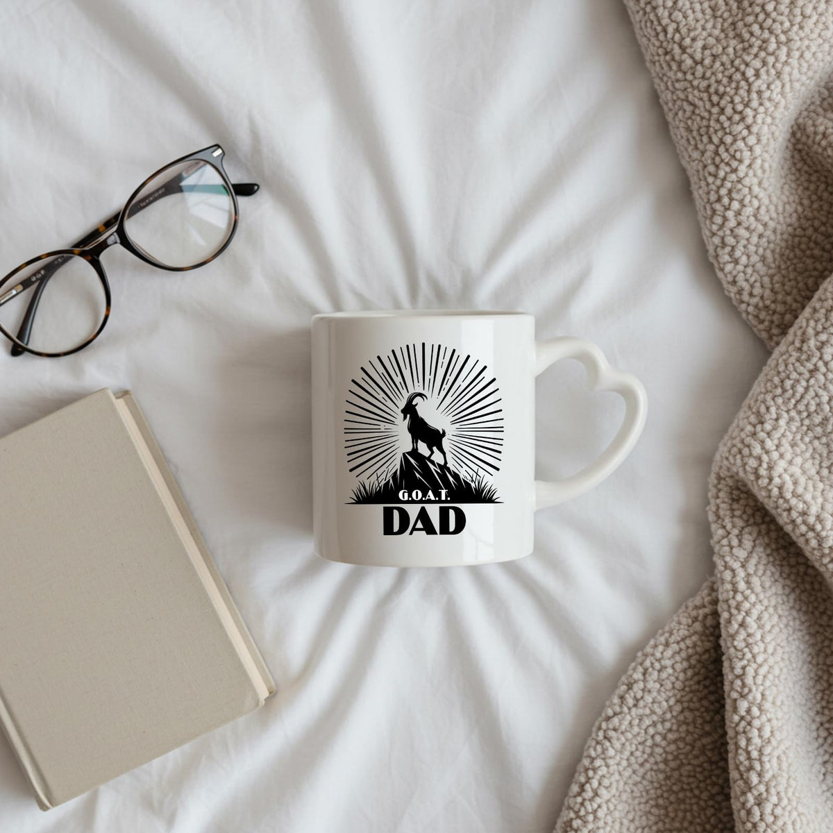 GOAT Dad heart-handle mug on a bed beside glasses and a book
