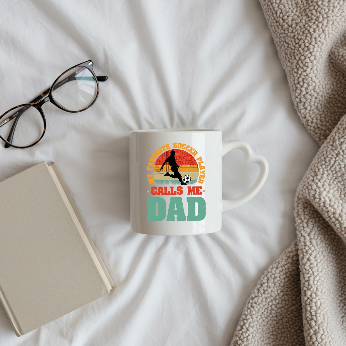 Soccer Dad heart-handle mug on a bed beside glasses and a book