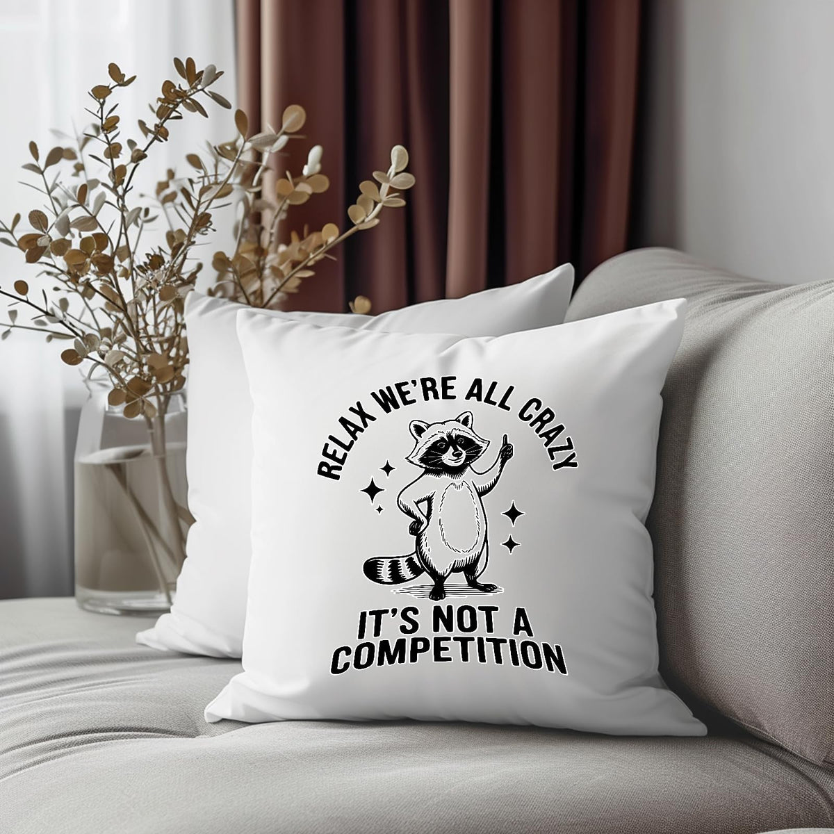 Relax We’re All Crazy raccoon cushion on a grey sofa beside dried flowers
