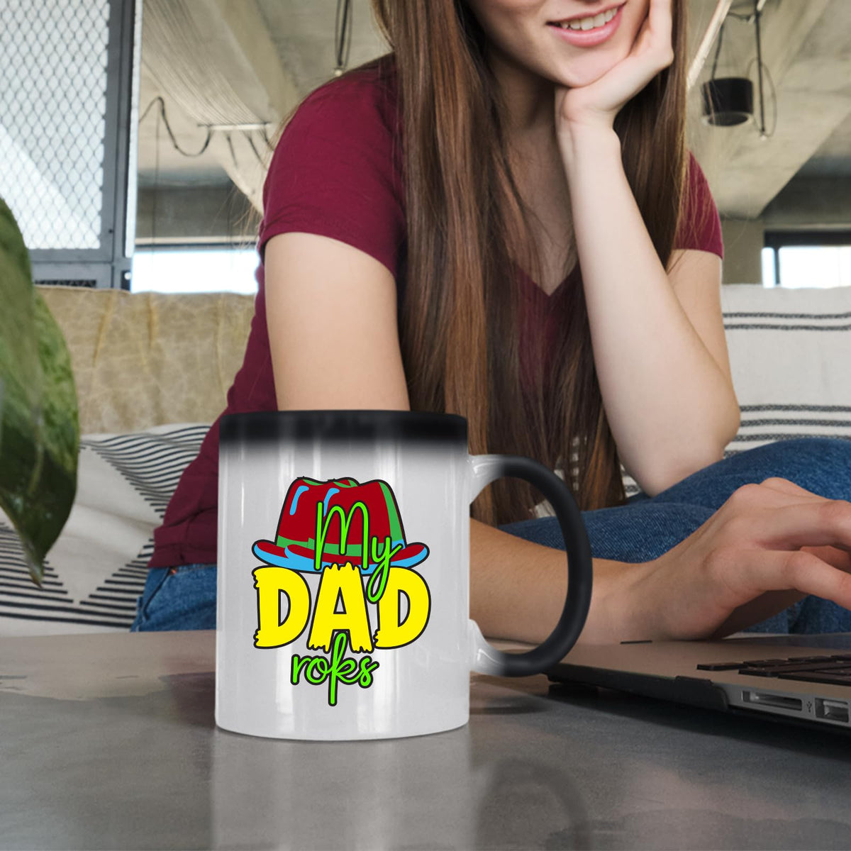 Woman at a laptop with a colour changing mug showing the colourful My Dad Rocks hat design