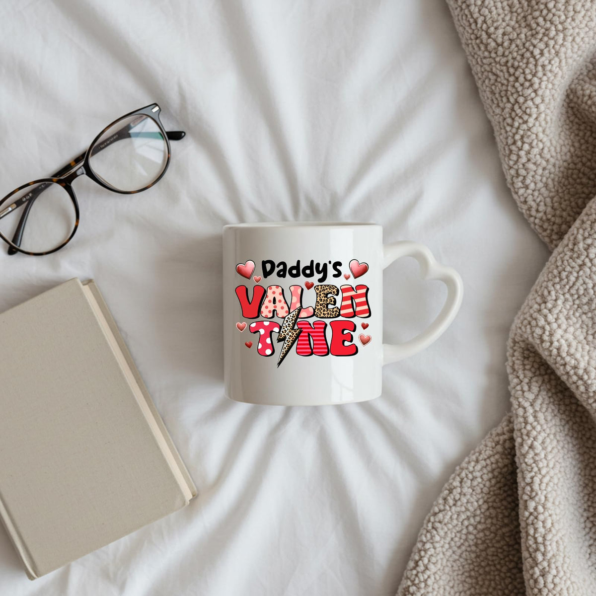 Daddy’s Valentine heart-handle mug on a bed beside glasses and a book