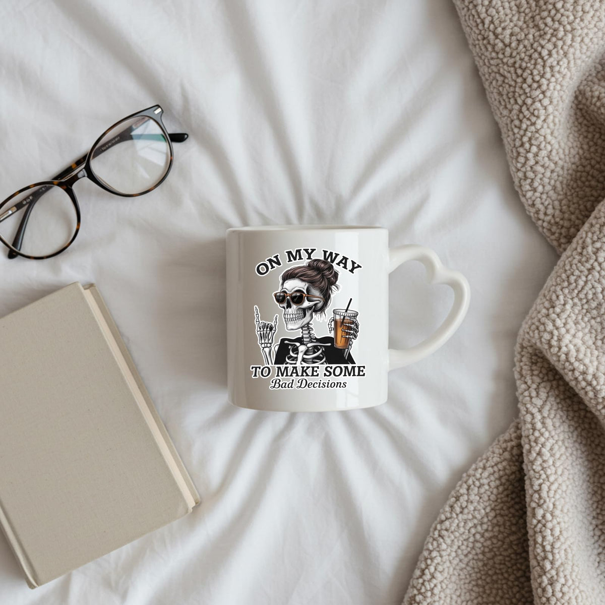 Bad Decisions skeleton heart-handle mug on a bed beside glasses and a book