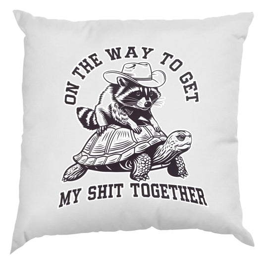 White cushion with a raccoon in a cowboy hat riding a tortoise and “On the way to get my shit together”
