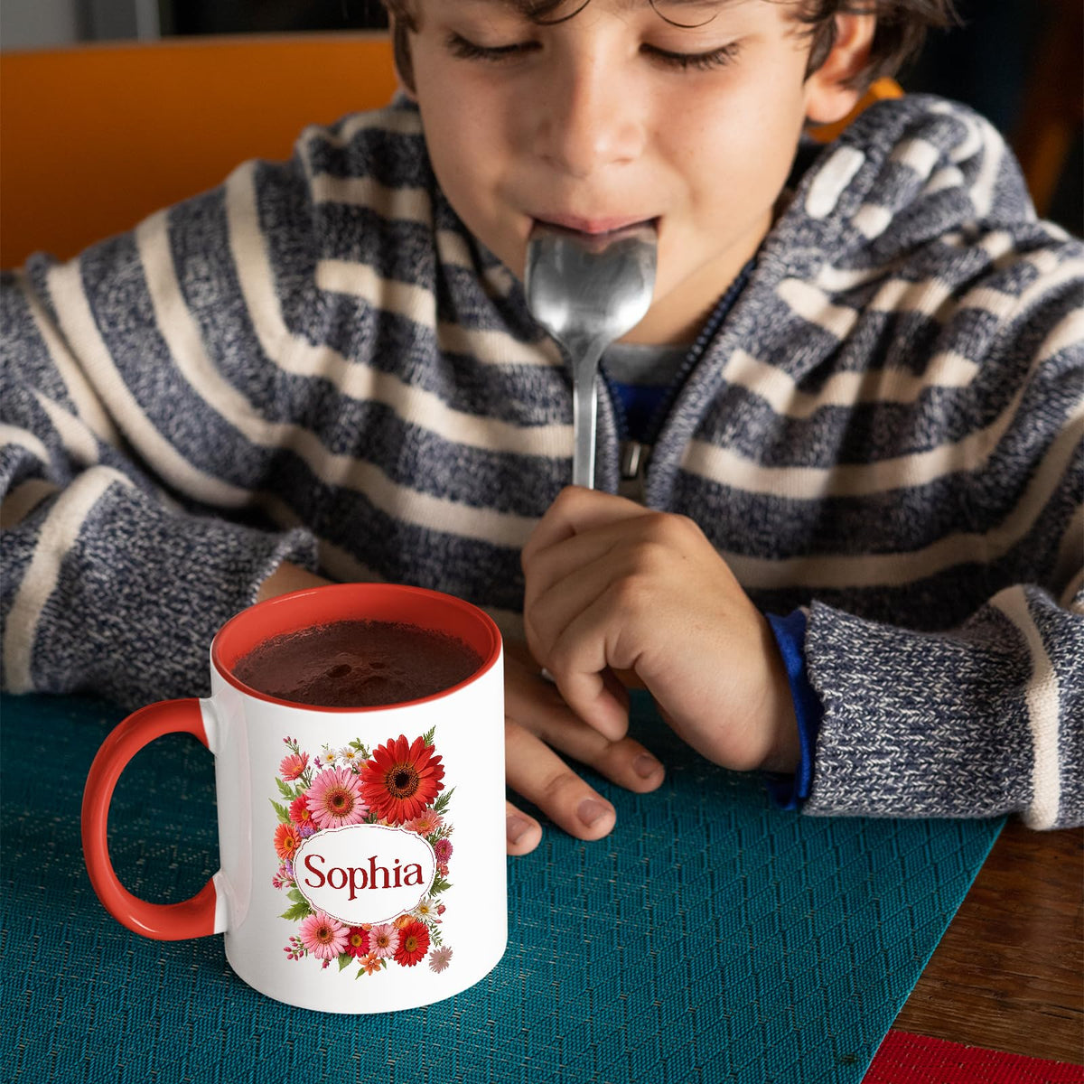 Boy with a spoon next to a red handle mug with a red gerbera wreath around an example name