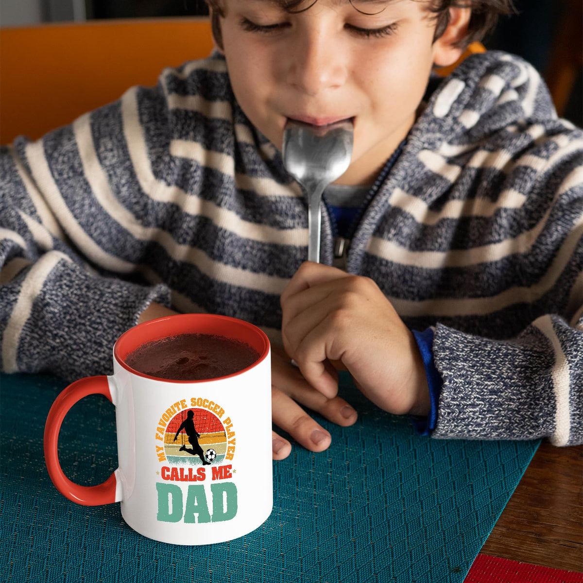 Child with a spoon next to the Soccer Dad mug with a red handle