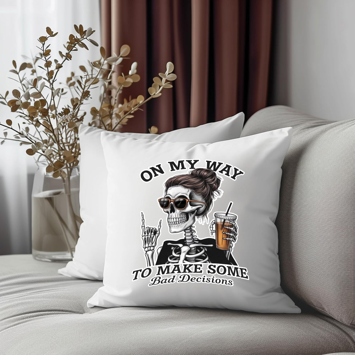 Bad Decisions skeleton cushion on a grey sofa beside dried flowers