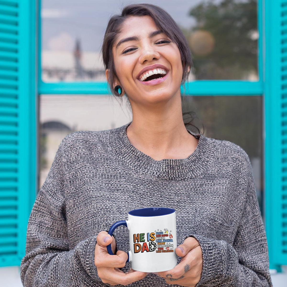 Smiling woman holding a dark blue handle mug with the He Is Dad Bible verse design