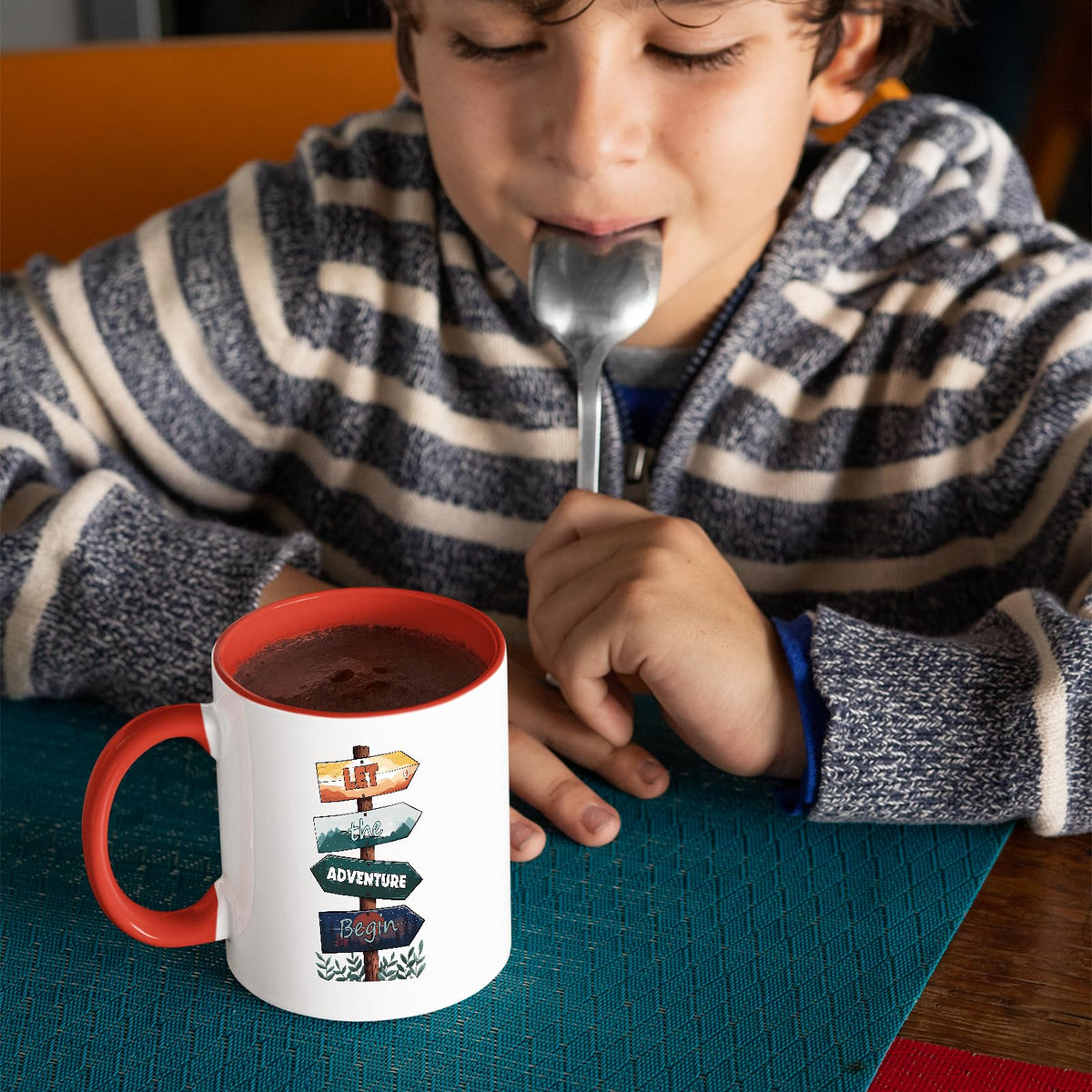 Child with a spoon next to the Let the Adventure Begin mug with a red handle