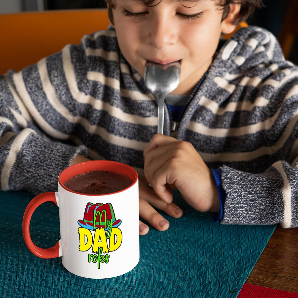 Boy with a spoon next to a red handle mug with the colourful My Dad Rocks hat design