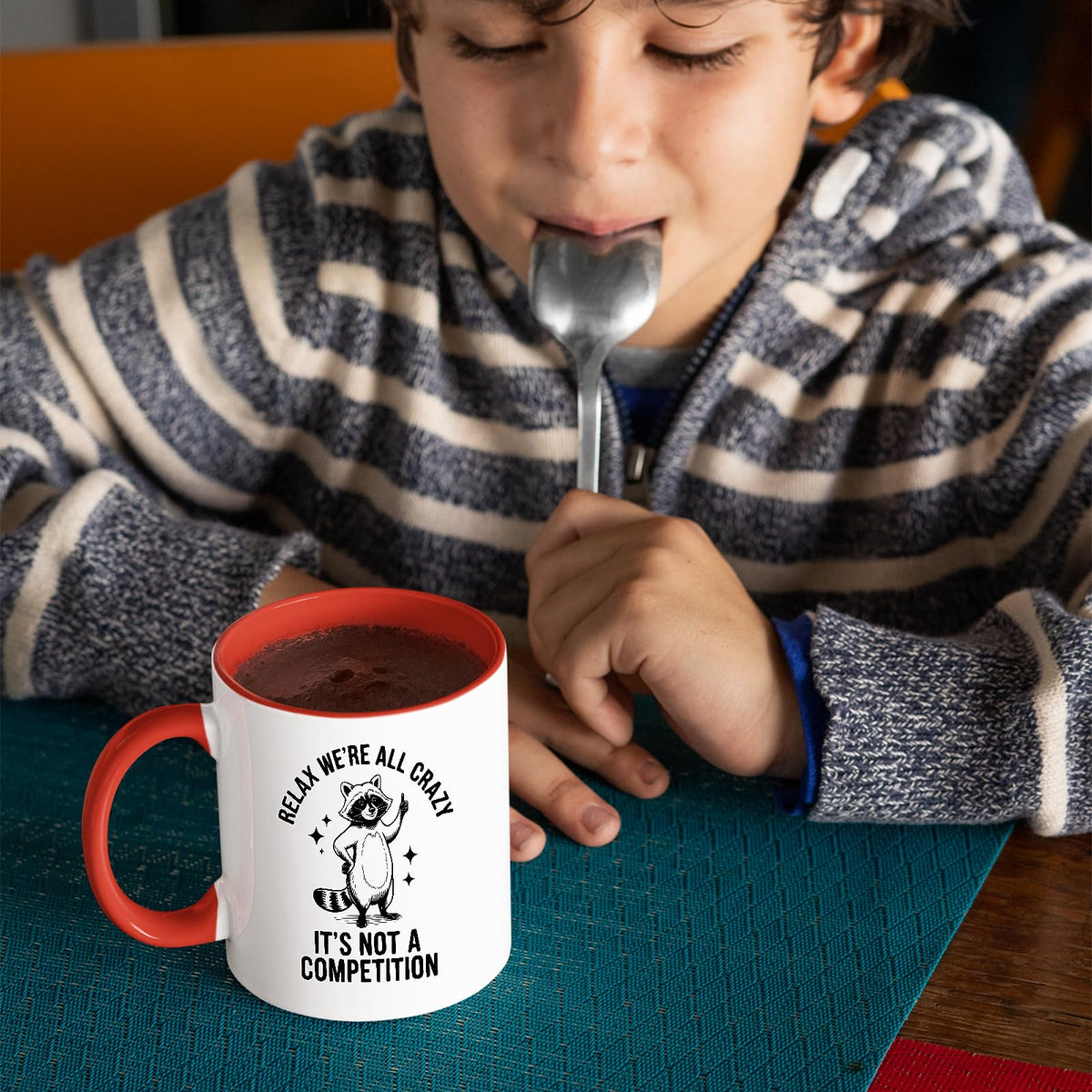 Child with a spoon next to the Relax We’re All Crazy raccoon mug with a red handle