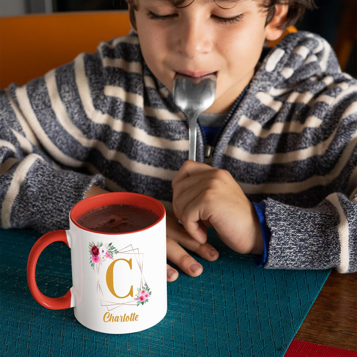 Boy with a spoon next to a red handle mug with a gold initial in a floral frame and an example name
