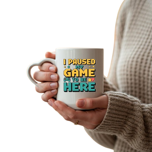 Hands holding the heart-handle mug with retro “I Paused My Game to Be Here” lettering
