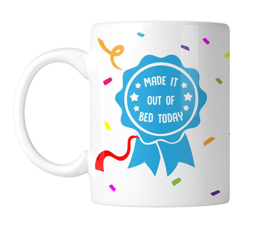 White mug with a blue award rosette that reads “Made It Out of Bed Today”, handle on the left